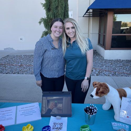 Two smiling women, possibly veterinarians, stand behind a table with informational materials, pens, and a stuffed dog. They are outside, next to a building with a window and a blue awning. A tablet displaying a QR code is on the table, alongside various flyers and colorful bracelets.