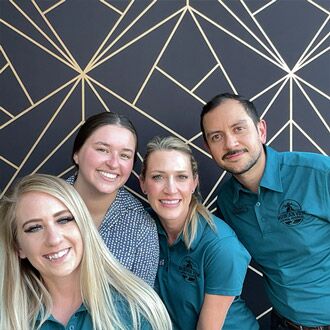 Four people are posing in front of a geometric-patterned wall. Three women and one man, presumably veterinarians, are smiling and all are wearing teal shirts with embroidered logos, except for one woman in a polka-dot blouse. They appear to be cheerful and friendly.